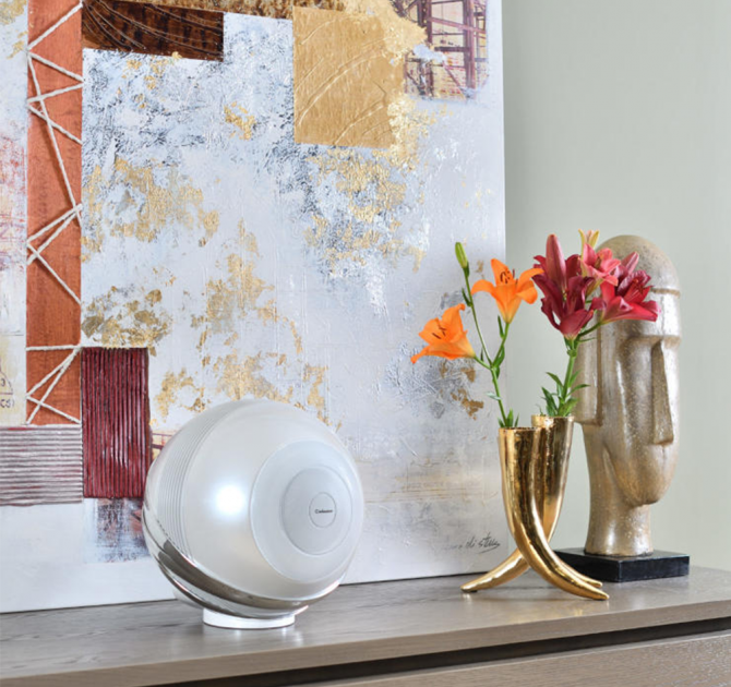 Cabasse Pearl Loudspeaker in white on a sideboard with flowers and an abstract picture behind.