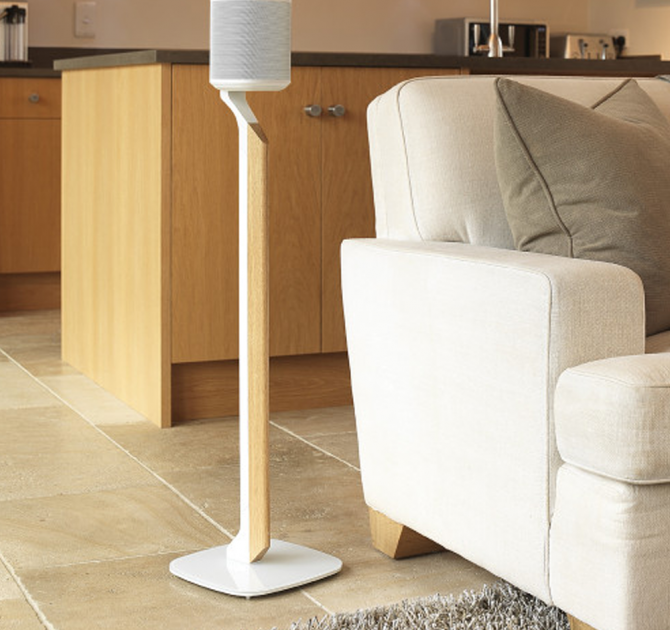 Flexson Premium Floor Stand One/Play1 Blk x1 - white with speaker on a tiled floor beside a sofa with a kitchen area in the background.