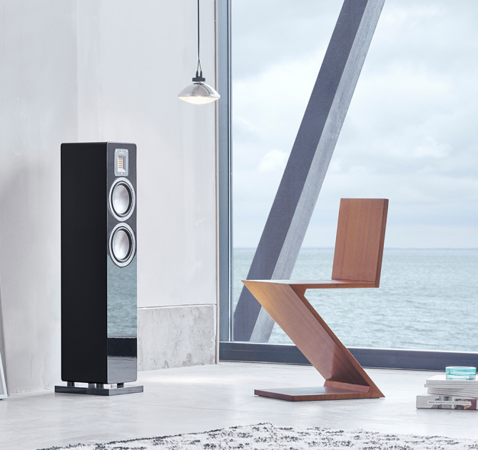 Audiovector QR3 in paino black beside a window with a seaview and a geometric style chair in front of it.
