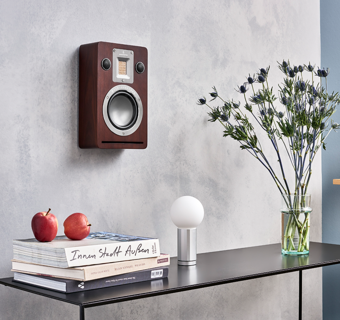 Audiovector QR Wall in dark walnut on a wall above a table with books, a lamp and flowers on