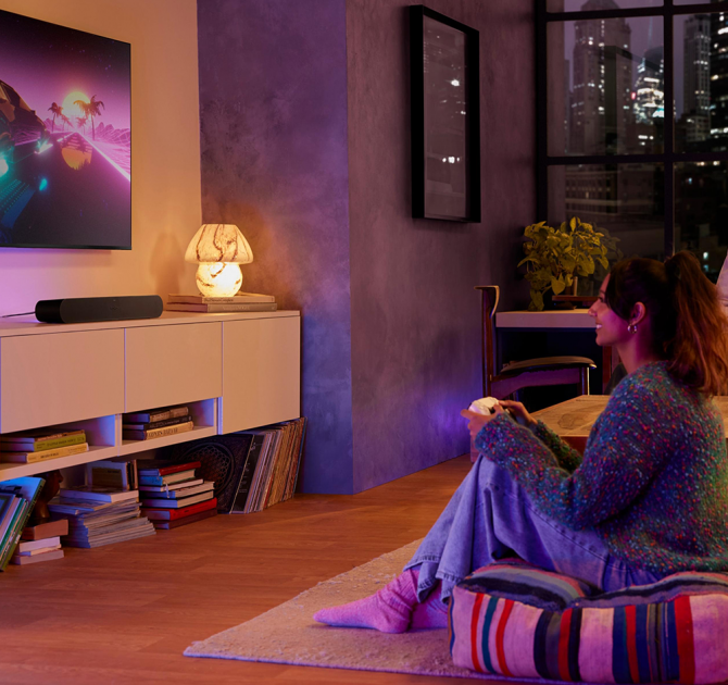 Sonos Ray Smart Soundbar in black on top of a tv stand with a wall mounted tv.  There's a woman sitting on the floor holding a gaming controller.