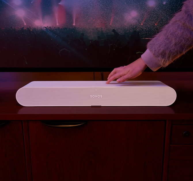 Sonos Ray Smart Soundbar in white in front of a tv with a hand touching the control on the top.
