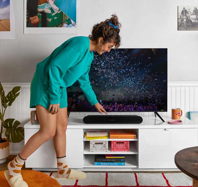 Sonos Ray Smart Soundbar in black on a tv stand with a woman bent over it pressing a control