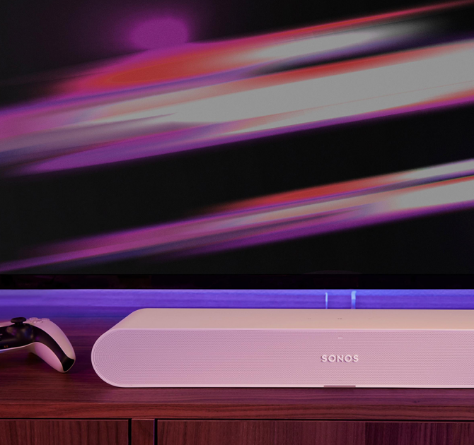 Sonos Ray Smart Soundbar in white on top of a sideboard with a gaming controller to the left.  There's a colourful tv screen in the background.
