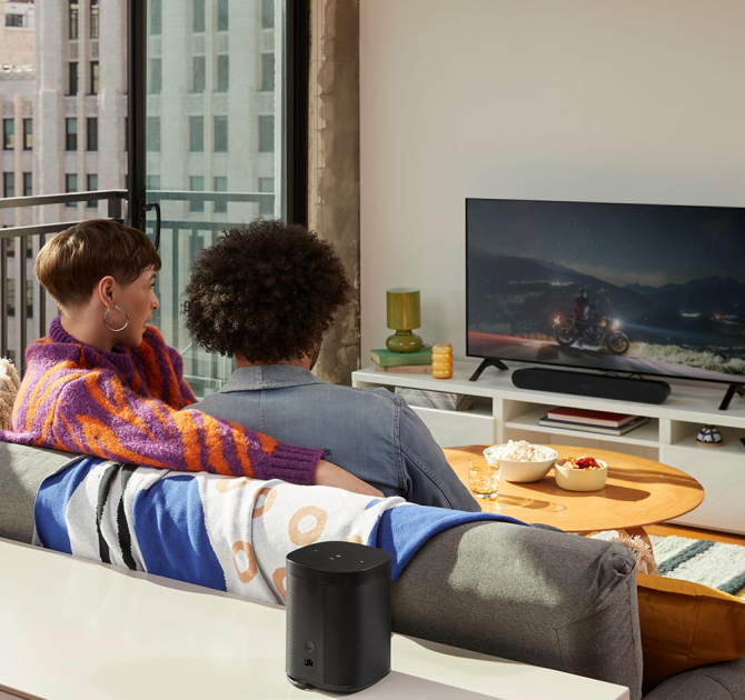 Sonos Ray Smart Soundbar in black on a tv stand with a couple in the foreground sitting on a sofa