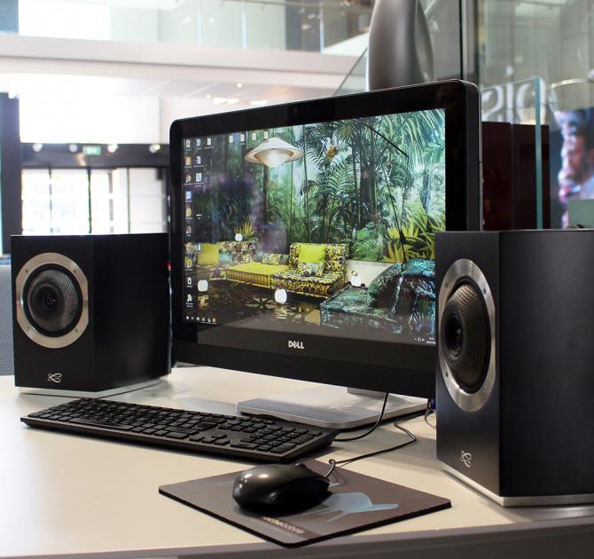 A pair of Cabasse Rialto Loudspeakers in black either side of a computer screen with a keyboard on the desk too.