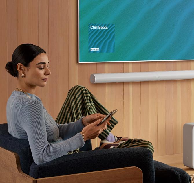 SONOS Arc Ultra Smart Soundbar in white on a wall underneath a tv with a woman sitting in a chair to the left of the image.