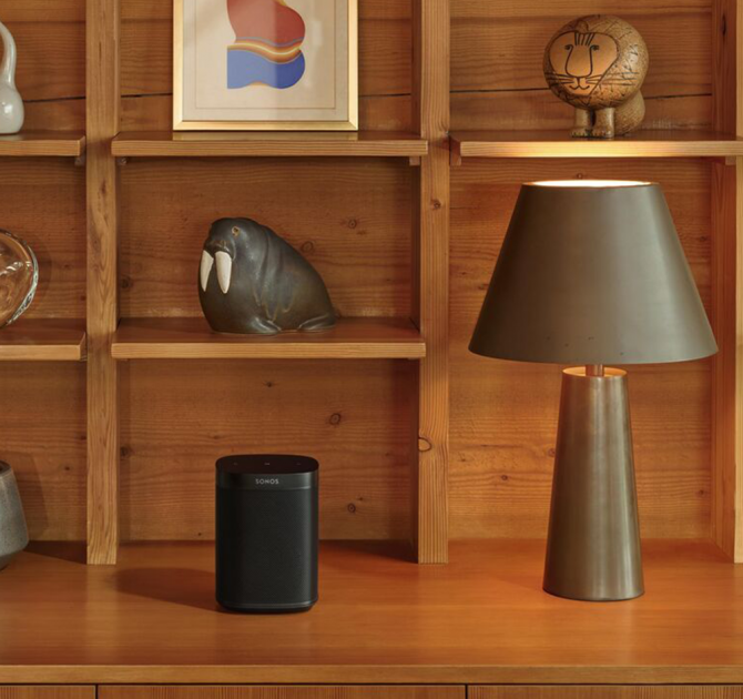 SONOS One SL Black on a wooden side board with rectangular section shelves holding a walrus ornament in one section, a lion ornament in another and an abstract red and blue picture in another.  There's a lamp beside the speaker.