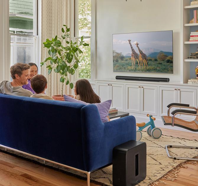 Sonos Sub Gen 4 in black beside a blue sofa with a family sat on it watching giraffes on tv.