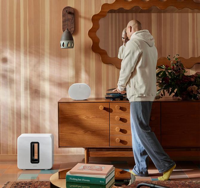 Sonos Sub Gen 4 in white on the floor beside a wooden sideboard with a man stood by the white speaker that's on top of the furniture