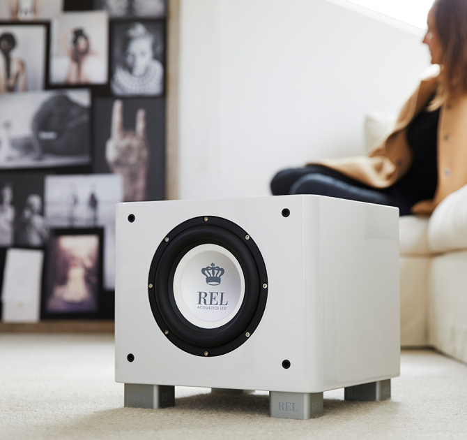 REL T/7x Sub-woofer in white on the floor beside a sofa with a photo wall in the background