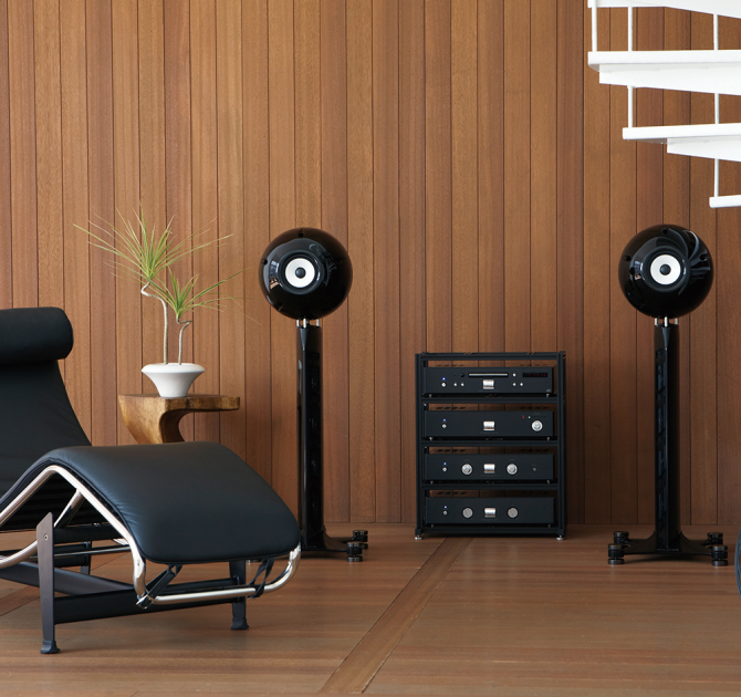Eclipse TD712zMK2 Speaker on Stand (Single) in black with a hifi system on shelving and a chair for lounging on.