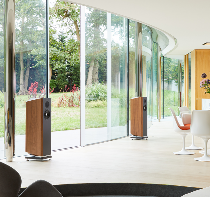 A pair of Kudos Titan 606 speakers with grill off in a large open plan living room in front of glass doors that open onto a garden.