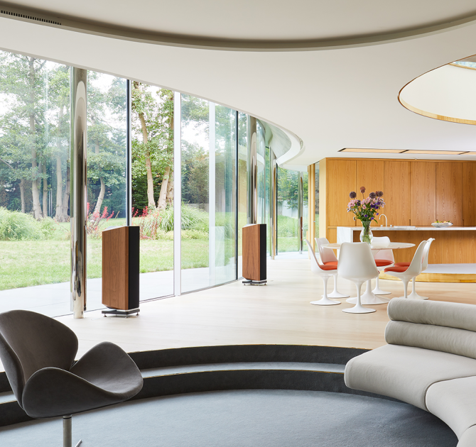 A pair of Kudos Titan 606 speakers with grill on in a large open plan living room in front of glass doors that open onto a garden.