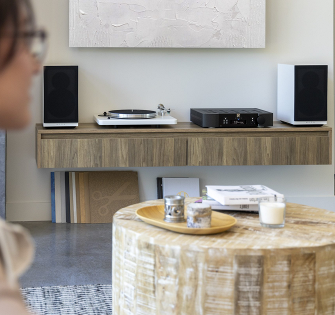 A pair of Moon Voice 22 Loudspeakers in white with grille on standing on a wooden sideboard.  There's a low wooden table in the middle of the image and a woman in the foreground.
