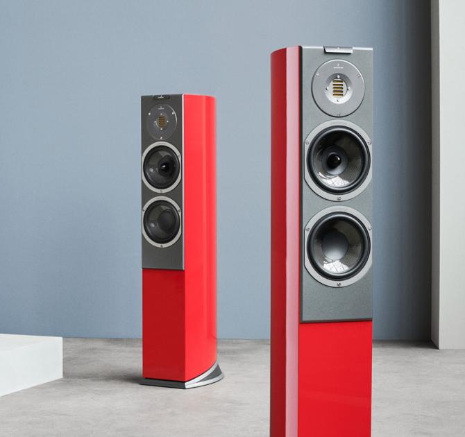 A pair of tall, slim red speakers in a room with a grey wall and a beige coloured floor.