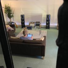 The ripcaster 360 speakers in Linn Heritage which is a dark blue colour.  There's a woman sitting on a brown sofa holding an iPad with her back to us.  Other HiFi equipment is between the two speakers.