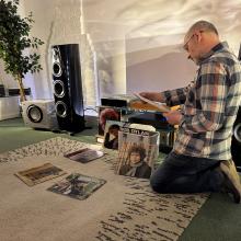 A man kneeling while reading the sleeve notes of an album.  there are Bob Dylan albums on the floor and stacked.  to the left of the image is the ripcaster 360 speaker in the Heritage colour.