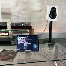 A white Linn 119 speaker at the ripcaster showroom.  It's on a stand beside an LP12 with an iPad in the foreground showing what's playing.