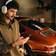 Sonos Ace Headphones on the head of a man who is looking at his phone.  In the background is a red porche