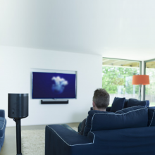 Flexson Adjustable Floor Stand One/Play1 with speaker and the view of a man from behind, sitting on a navy sofa watching a wall mounted television with an orange standard lamp in the corner of the room in front of a large double-aspect window.