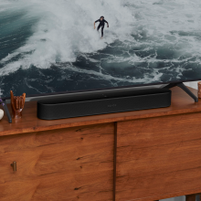 SONOS Beam (Gen 2) in black on top of a tv unit.  the tv has a picture of a surfer in a black wetsuit riding a wave.
