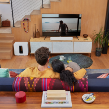 SONOS Beam (Gen 2) in white on top of a tv cabinet.  There is a Sonos Sub in white on the floor.  the tv is showing a man with his back to us looking out of a large window onto a very plain landscape.  there's a man and woman in the foreground with their back to us sitting close on a sofa.