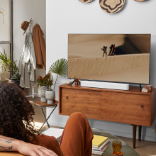SONOS Beam (Gen 2) in white on a wooden tv cabinet.  there's a woman in the foreground with a man's arm along the sofa behind her.  the tv cabinet has a small table beside it with two vases containing foilage.  There's a hat stand to the side of the tv.  the tv is showing two people walking on sand dunes.
