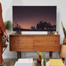 SONOS Beam (Gen 2) in white on top of a wooden tv unit.  there's a woman standing with the tv remote in her hand.  She's on the right of the image and only part of her can be seen.  the tv is showing something that involves a sunset with a line of trees in silhouette.