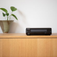 Melco D100 Compact Disc Drive in black on top of a wooden side-board with a plant to the left.