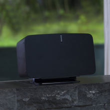 Flexson Desk Stand with a black Sonos Play 5 on it on a dark brick windowsill in front of a window.
