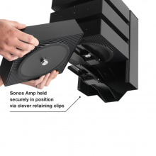 Flexson Dock x4 Amp Black x1 with hands holding an amp about to be put into the empty slot.  Annotated "Sonos Amp held securely in position via clever retaining clips."