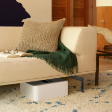 SONOS Sub in white laying down under a sofa that has an open book and a pair of glasses laying on it.