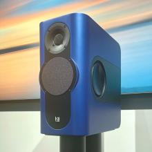A blue rectangular speaker on a black stand with a piece of artwork in the background.