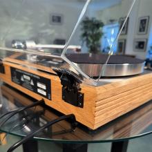 Linn Majik MM LP12 Fluted turntable in oak in the ripcaster showroom with sofas and speakers in the background along with other HiFi equipment.  This image shows a back corner with the hinge in focus