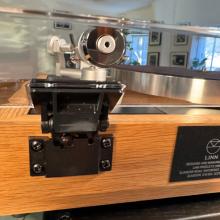Linn Majik MM LP12 Fluted turntable in oak in the ripcaster showroom with sofas and speakers in the background along with other HiFi equipment.  This image is a close-up of a back hinge and the back of the tonearm.