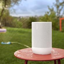 SONOS Move 2 Loudspeaker in white on a red table outside with a sprinkler.  The speaker has water droplets on it.