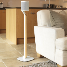 Flexson Premium Floor Stand One/Play1 Blk x1 - white with speaker on a tiled floor beside a sofa with a kitchen area in the background.