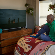 Sonos Ray Smart Soundbar in black in front of a tv with a man sitting at the end of a bed with a colourful throw on it.