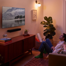 Sonos Ray Smart Soundbar in black mounted on the wall with a tv screen above.  There's a couple, one on the sofa and one sitting on the floor in front of the sofa.