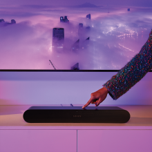 Sonos Ray Smart Soundbar in black in front of a tv with a hand touching the control on the top.