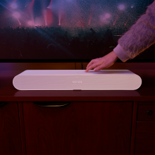 Sonos Ray Smart Soundbar in white in front of a tv with a hand touching the control on the top.