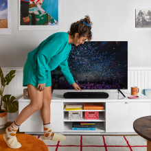 Sonos Ray Smart Soundbar in black on a tv stand with a woman bent over it pressing a control