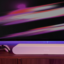Sonos Ray Smart Soundbar in white on top of a sideboard with a gaming controller to the left.  There's a colourful tv screen in the background.