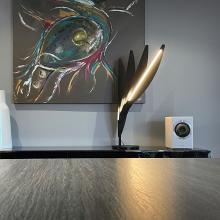 A Cabasse Rialto Loudspeaker on top of a unit with a modern lamp beside it and a piece of abstract art on the wall behind.
