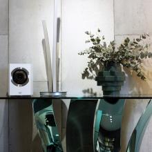 A Cabasse Rialto Loudspeaker in white on top of a glass table with a vase of eucalyptus on the right.
