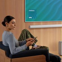 SONOS Arc Ultra Smart Soundbar in white on a wall underneath a tv with a woman sitting in a chair to the left of the image.