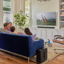 Sonos Sub Gen 4 in black beside a blue sofa with a family sat on it watching giraffes on tv.