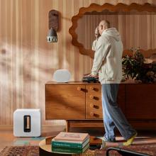 Sonos Sub Gen 4 in white on the floor beside a wooden sideboard with a man stood by the white speaker that's on top of the furniture