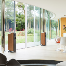 A pair of Kudos Titan 606 speakers with grill off in a large open plan living room in front of glass doors that open onto a garden.